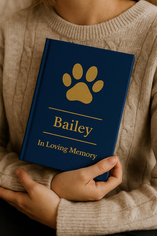 Person gently holding a personalized pet memorial journal close to their chest, navy blue matte hardcover with gold paw print and the name Bella between gold divider lines, comforting remembrance keepsake for mourning pet parents.