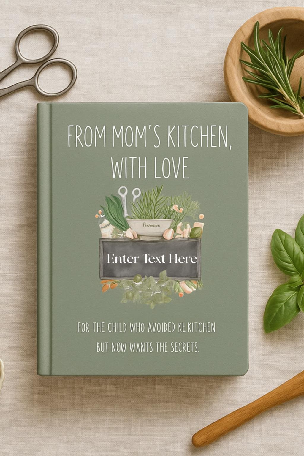Flat lay of sage green recipe legacy notebook titled ‘From Mom’s Kitchen, With Love.’ Styled with rustic kitchen tools including a wooden cutting board, scissors, knife, mortar and pestle, peppercorns, rosemary, and basil leaves. Inviting family cookbook gift idea.